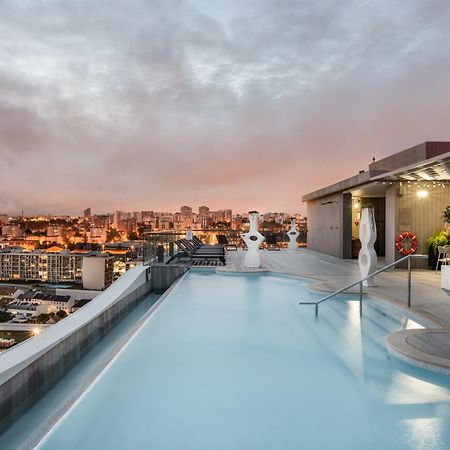 Отель Eurostars Universal Lisboa Экстерьер фото The photo shows a rooftop pool with a modern design, featuring clear blue water and a sleek edge that appears to blend into the skyline. In the background, there are city buildings illuminated by sunset colors, giving the scene a warm and inviting at