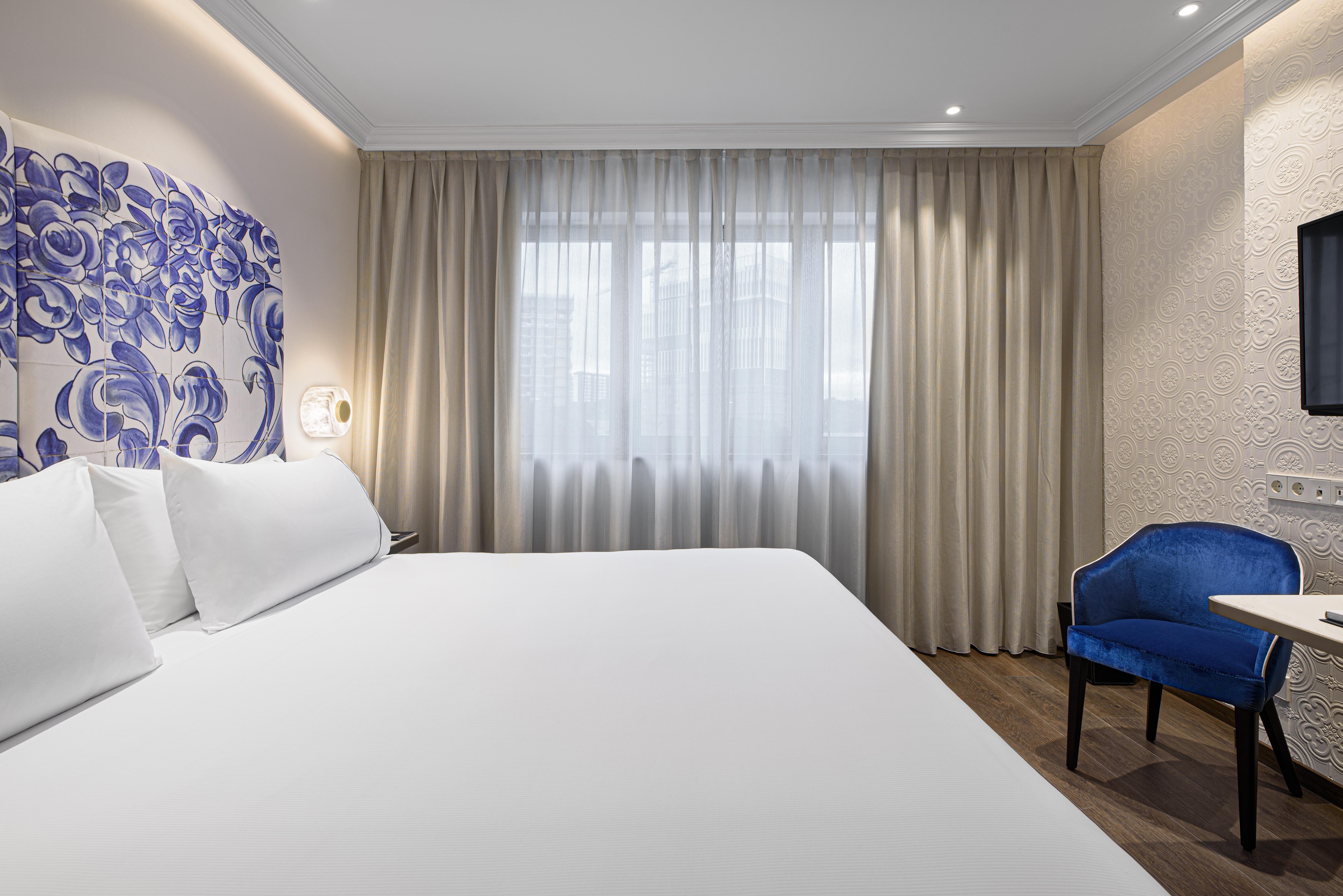 Отель Eurostars Universal Lisboa Экстерьер фото The photo shows a modern hotel room with a minimalistic design. It features a large bed with white linens and two pillows. Behind the bed, there is a decorative wall with a blue floral design. To the side, a window is covered with sheer curtains, all