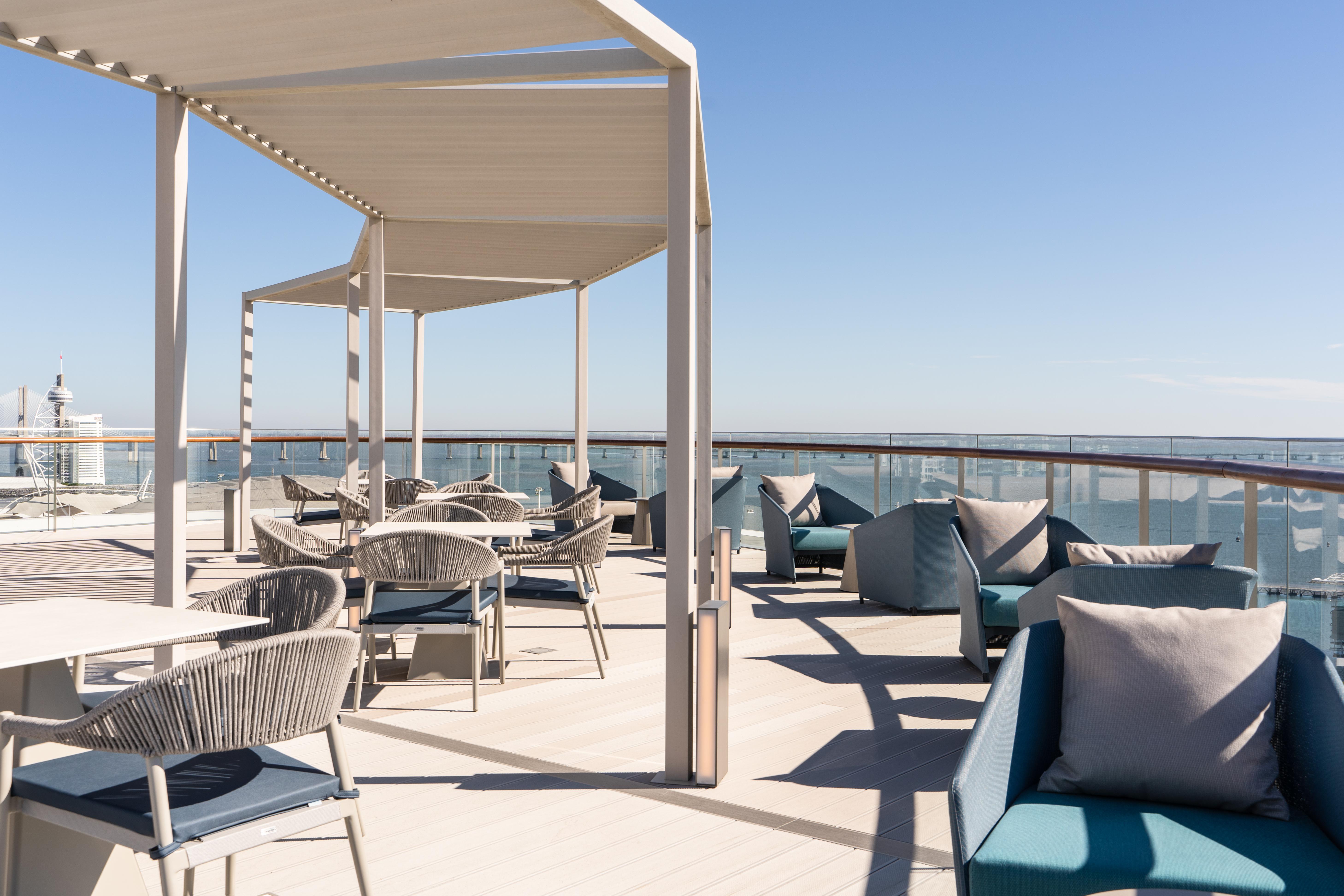 Отель Eurostars Universal Lisboa Экстерьер фото The photo shows a spacious outdoor seating area on a deck or terrace. There are several stylish chairs and tables arranged under a white canopy for shade. The furniture appears contemporary, with a mix of materials including woven textures. The backd