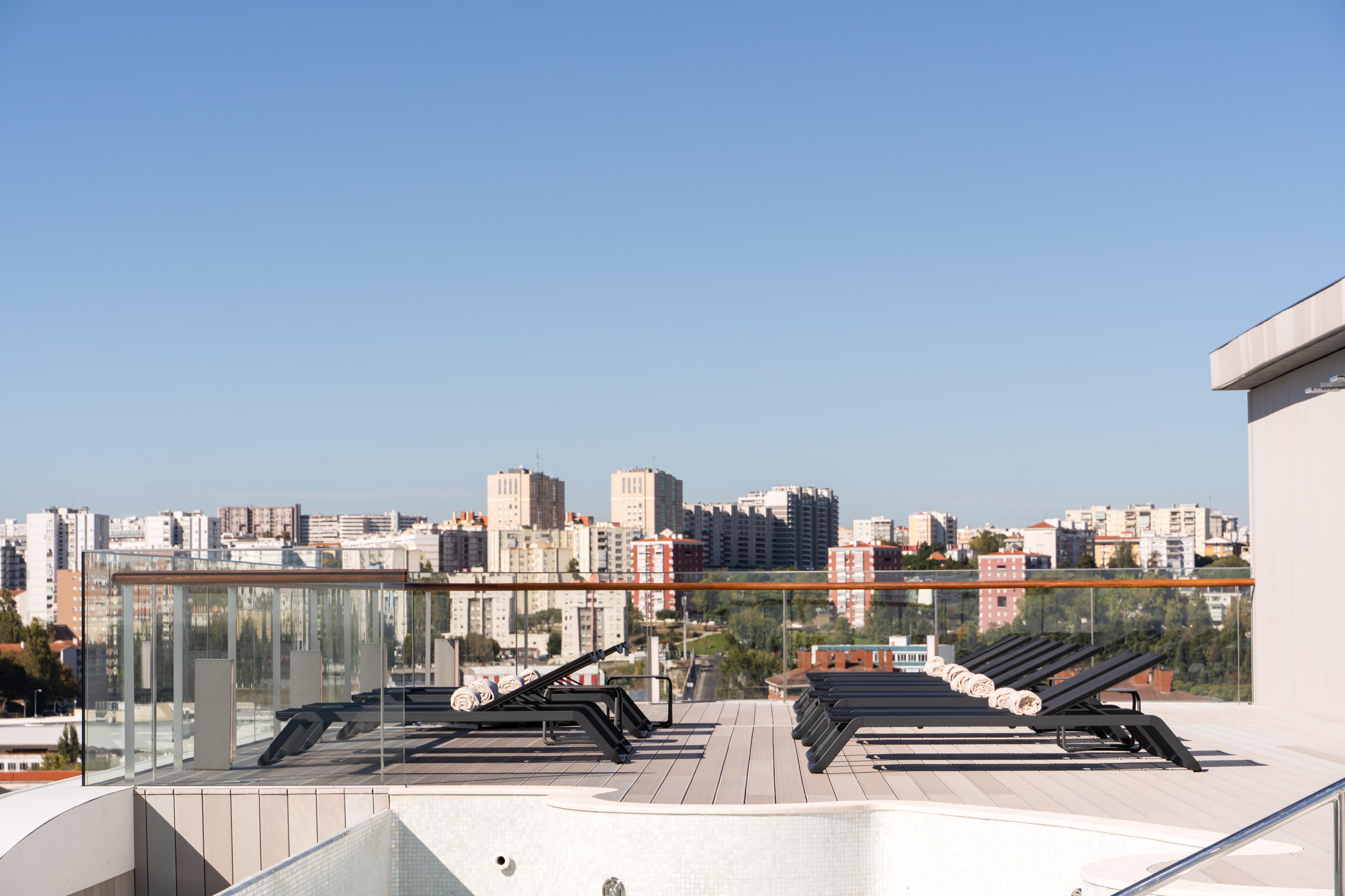 Отель Eurostars Universal Lisboa Экстерьер фото The image shows a rooftop setting with a clear blue sky. In the foreground, there are several lounge chairs arranged on a wooden deck. Beyond the deck, a transparent railing allows for an unobstructed view of a cityscape, which includes various build