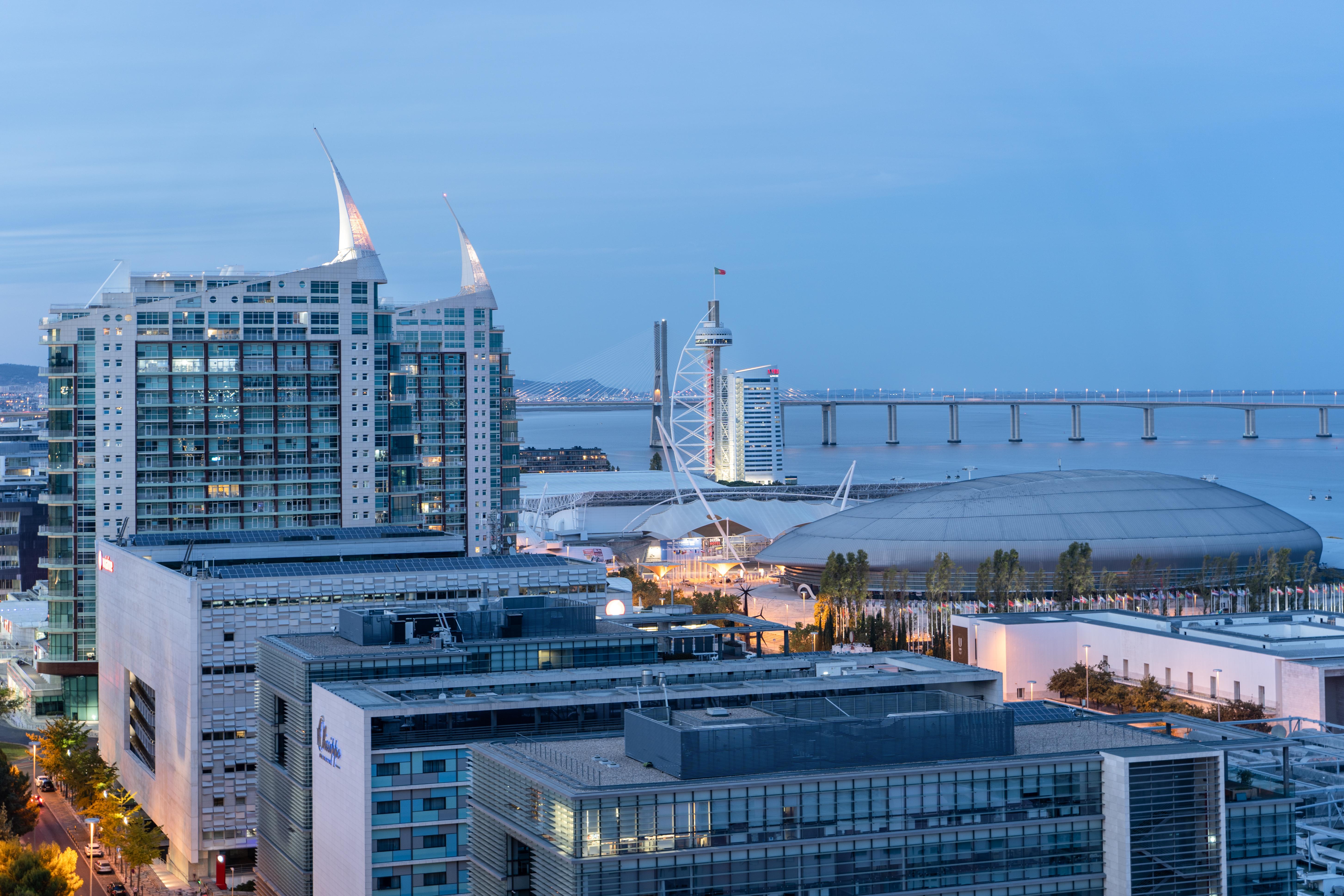 Отель Eurostars Universal Lisboa Экстерьер фото The photo depicts a modern urban landscape at dusk, showcasing a combination of tall buildings and structures. In the foreground, there's a cluster of contemporary architecture, including large glass buildings with a sleek design. In the background, 