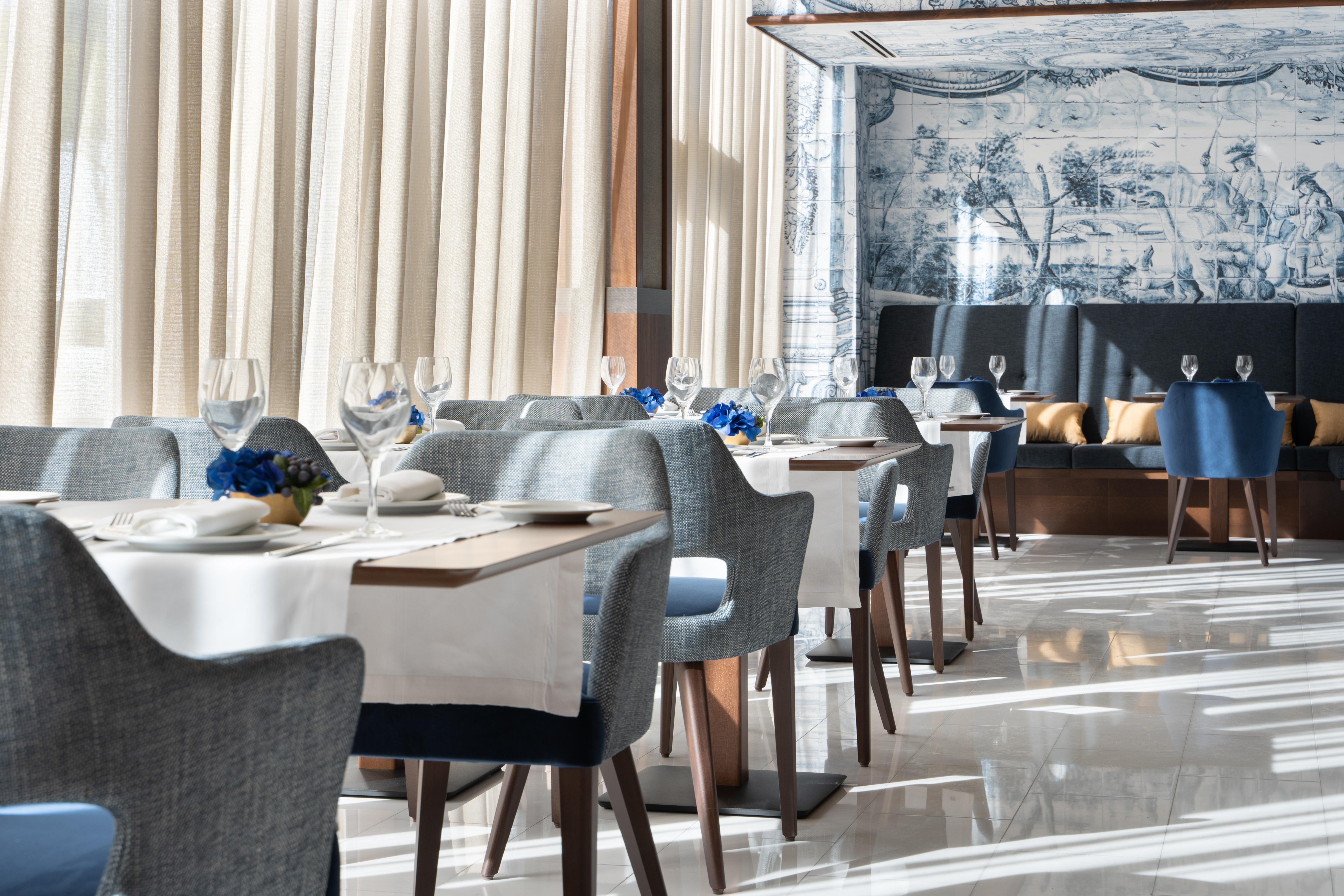 Отель Eurostars Universal Lisboa Экстерьер фото The photo shows an elegant restaurant interior. It features modern dining tables set with white tablecloths and neatly arranged tableware, including wine glasses. The chairs are upholstered in a soft gray fabric, and there are decorative elements lik