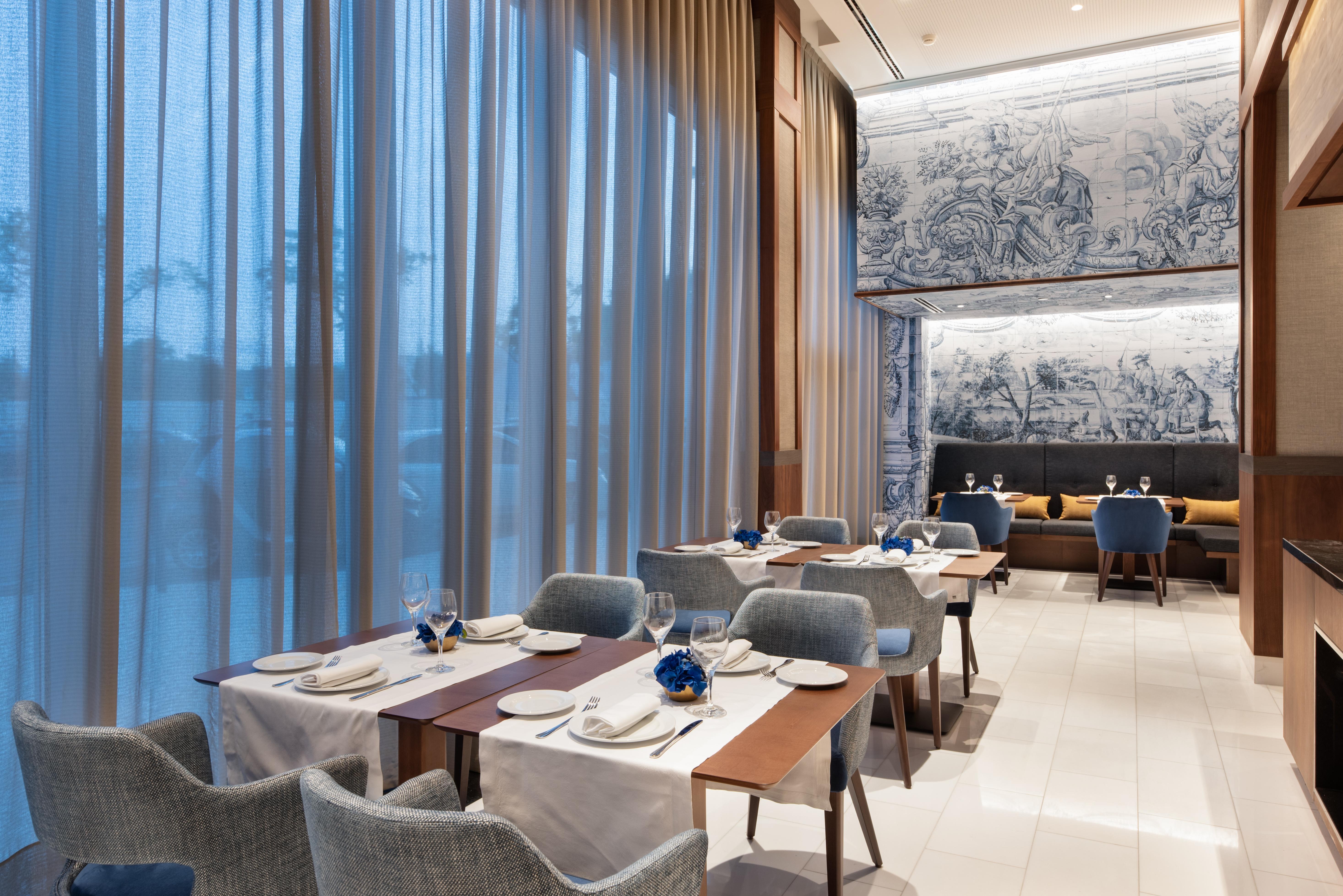 Отель Eurostars Universal Lisboa Экстерьер фото The photo shows a modern dining area in a restaurant. There are several tables set with white tablecloths, plates, glasses, and decorative elements. The chairs are upholstered in a soft blue fabric. Large windows draped with sheer curtains allow natu