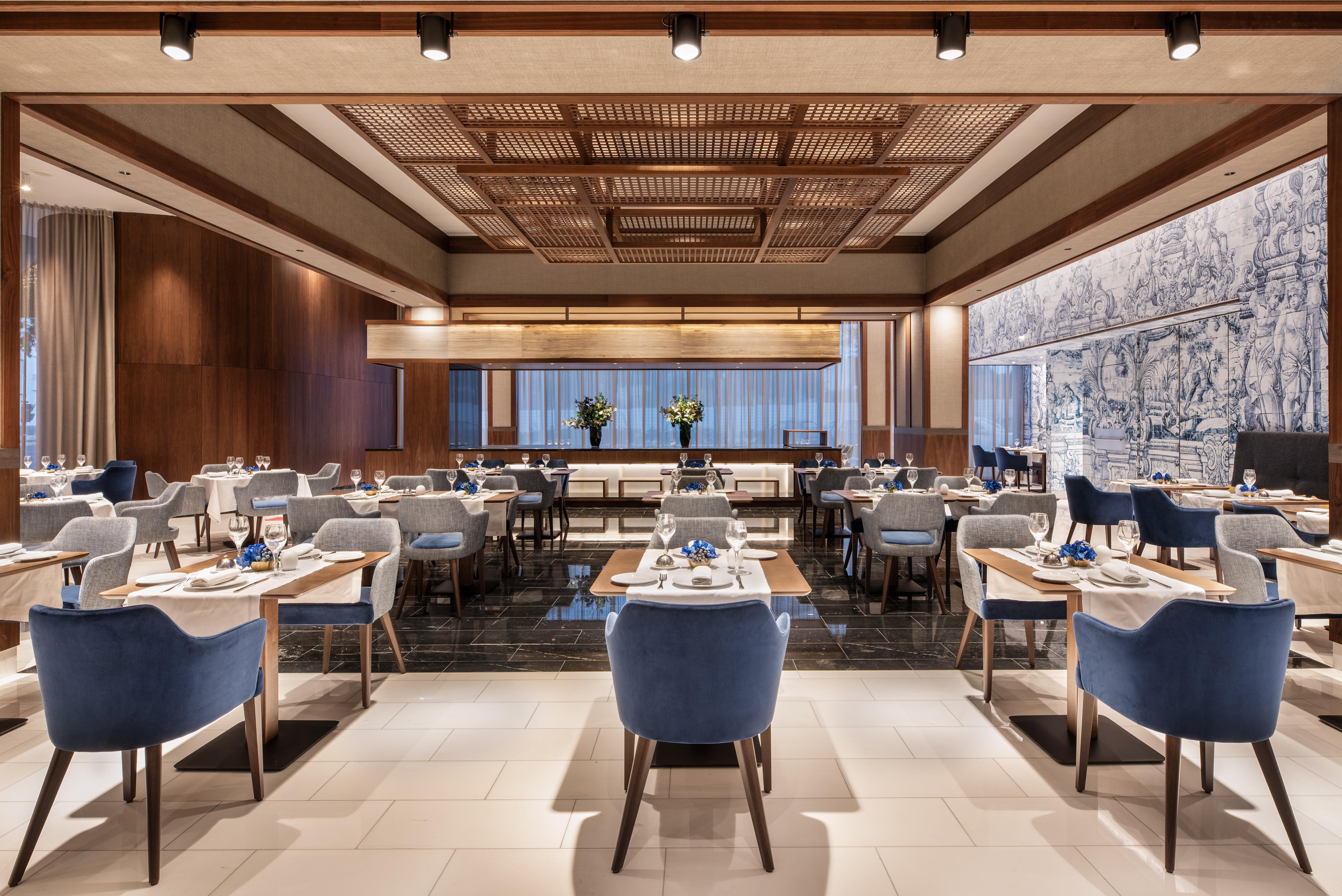 Отель Eurostars Universal Lisboa Экстерьер фото The photo shows an interior view of a modern restaurant. The space features a contemporary design with a warm color palette, including wood paneling and elegant blue upholstered chairs. There are several tables set for dining, with white tablecloths 