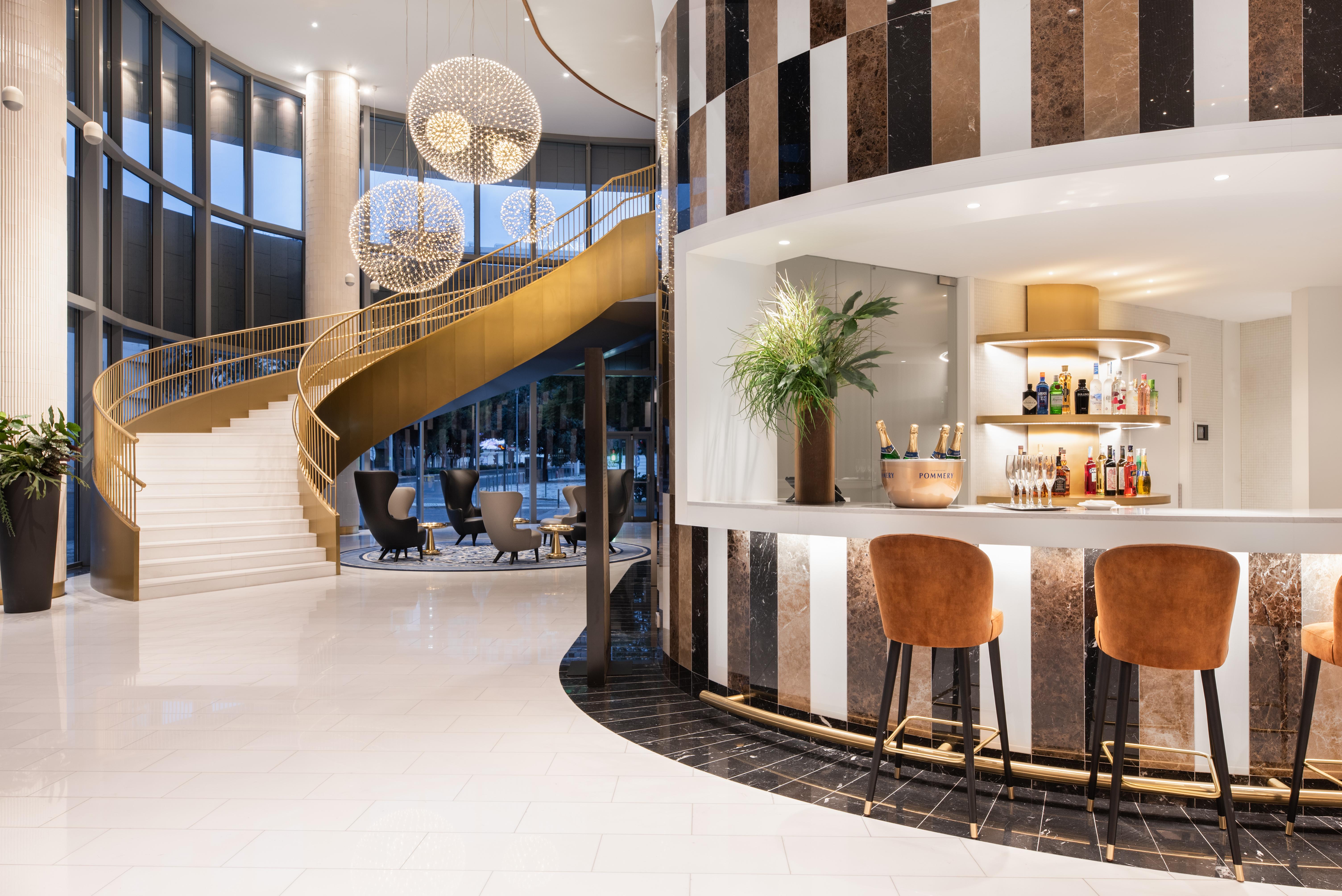 Отель Eurostars Universal Lisboa Экстерьер фото The photo depicts a stylish, modern interior space featuring a curved staircase with a golden railing. The staircase leads to an upper level and is illuminated by large pendant lights. To the right, there is a sleek bar area with a white countertop, 