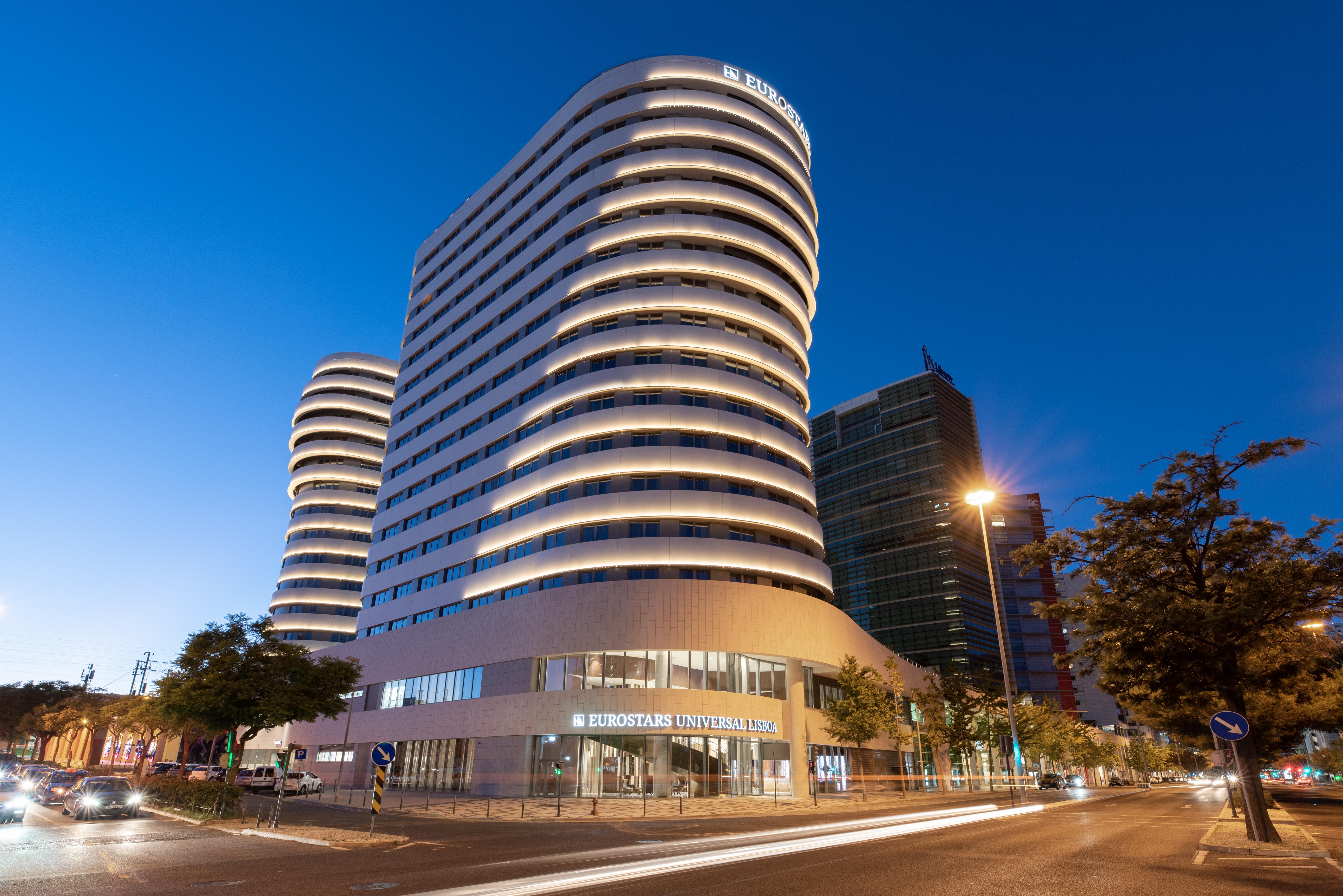 Отель Eurostars Universal Lisboa Экстерьер фото The photo shows a modern building complex featuring two distinct towers. The towers have a smooth, curved facade with horizontal lines that create a sleek, contemporary look. The building is illuminated with soft lighting that highlights its architec