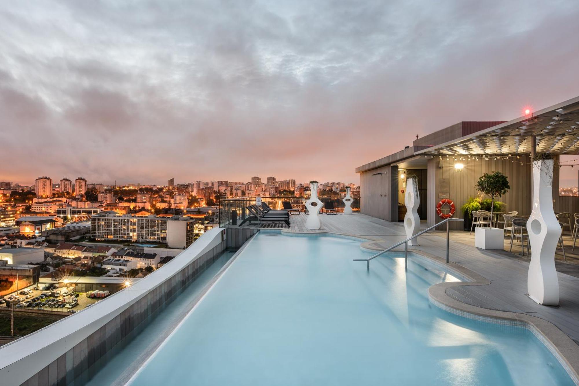 Отель Eurostars Universal Lisboa Экстерьер фото The photo shows a rooftop pool with a modern design, featuring clear blue water and a sleek edge that appears to blend into the skyline. In the background, there are city buildings illuminated by sunset colors, giving the scene a warm and inviting at