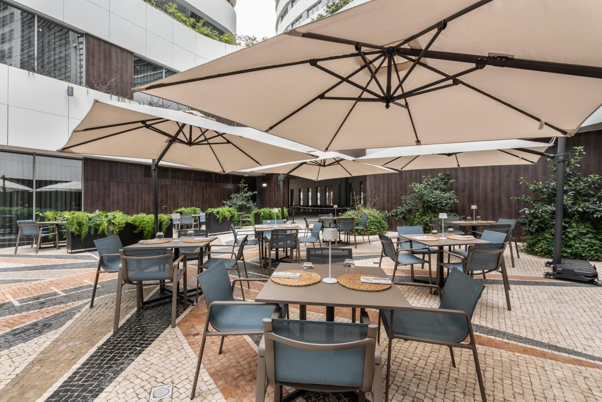 Отель Eurostars Universal Lisboa Экстерьер фото The photo shows an outdoor dining area featuring several tables and chairs set up beneath large umbrellas. The space is designed with a modern aesthetic, surrounded by greenery and potted plants. The flooring consists of patterned tiles, adding to th