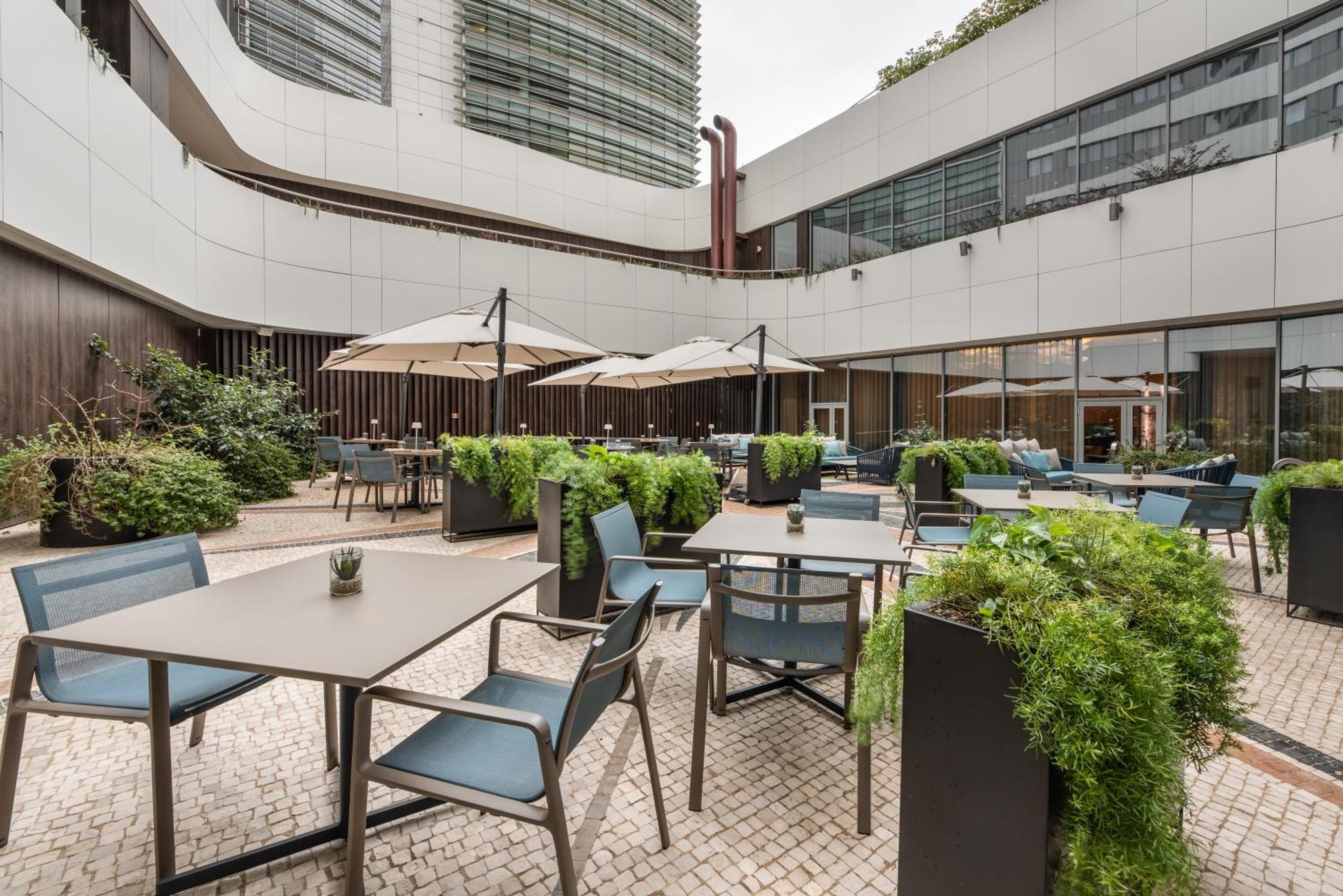 Отель Eurostars Universal Lisboa Экстерьер фото The image shows a modern outdoor seating area, likely part of a restaurant or cafe. It features several tables and chairs arranged neatly, with some tables covered by umbrellas for shade. Lush greenery surrounds the area, with planters filled with pl