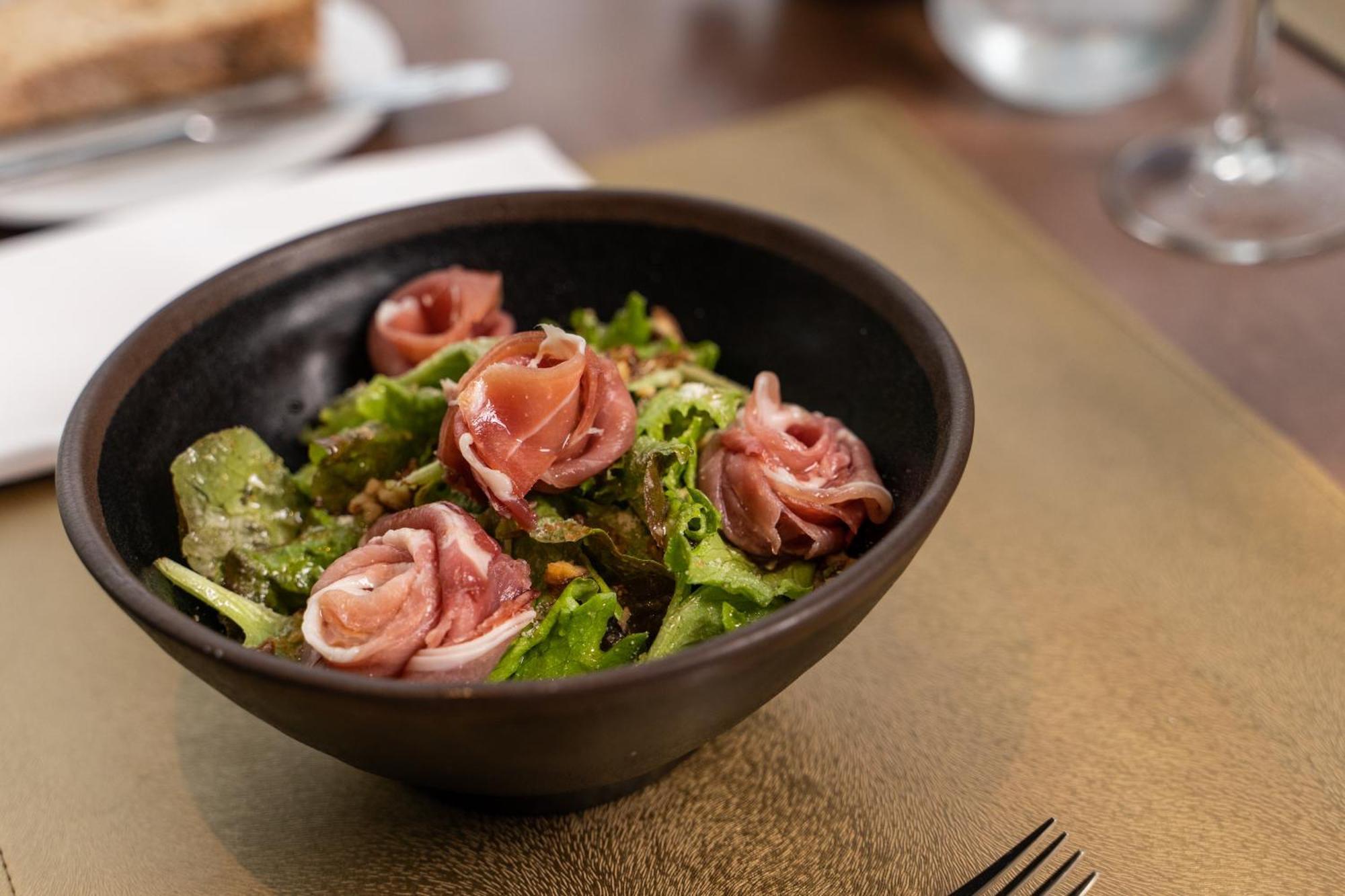 Отель Eurostars Universal Lisboa Экстерьер фото The photo shows a bowl of salad with a mix of fresh greens, topped with rolled slices of cured meat, likely ham. The salad has a vibrant color, featuring leafy greens and the pinkish hue of the meat. The bowl is dark and round, with a simple, elegant