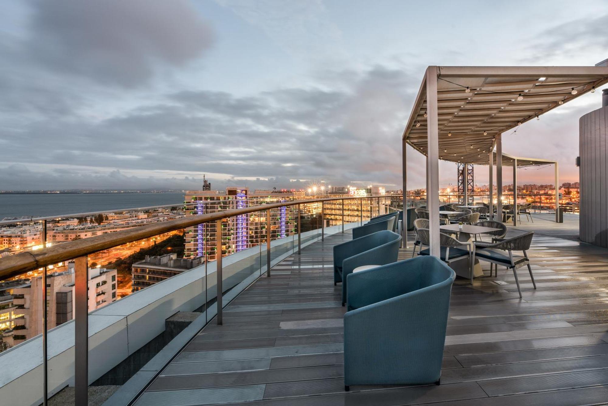 Отель Eurostars Universal Lisboa Экстерьер фото The photo shows a rooftop terrace with a modern design. There are several blue chairs grouped around small tables, ideal for relaxation or dining. The terrace has a covered area which provides shade, and it overlooks a cityscape with buildings and a 