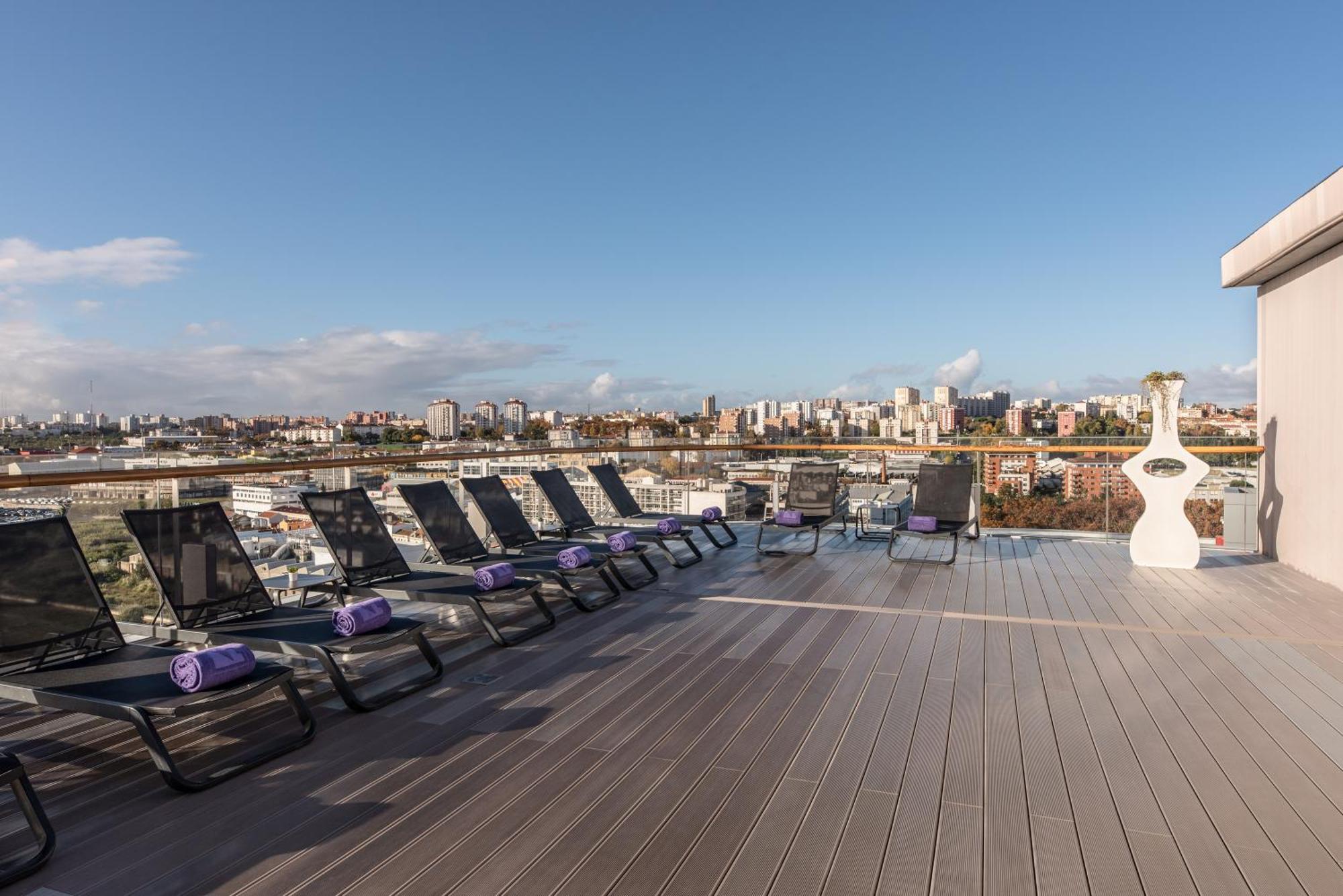 Отель Eurostars Universal Lisboa Экстерьер фото The photo shows a rooftop terrace with several lounge chairs arranged in a row. Each chair is equipped with a purple towel. The terrace has a wooden deck and offers a panoramic view of a cityscape with various buildings in the background. The sky is 