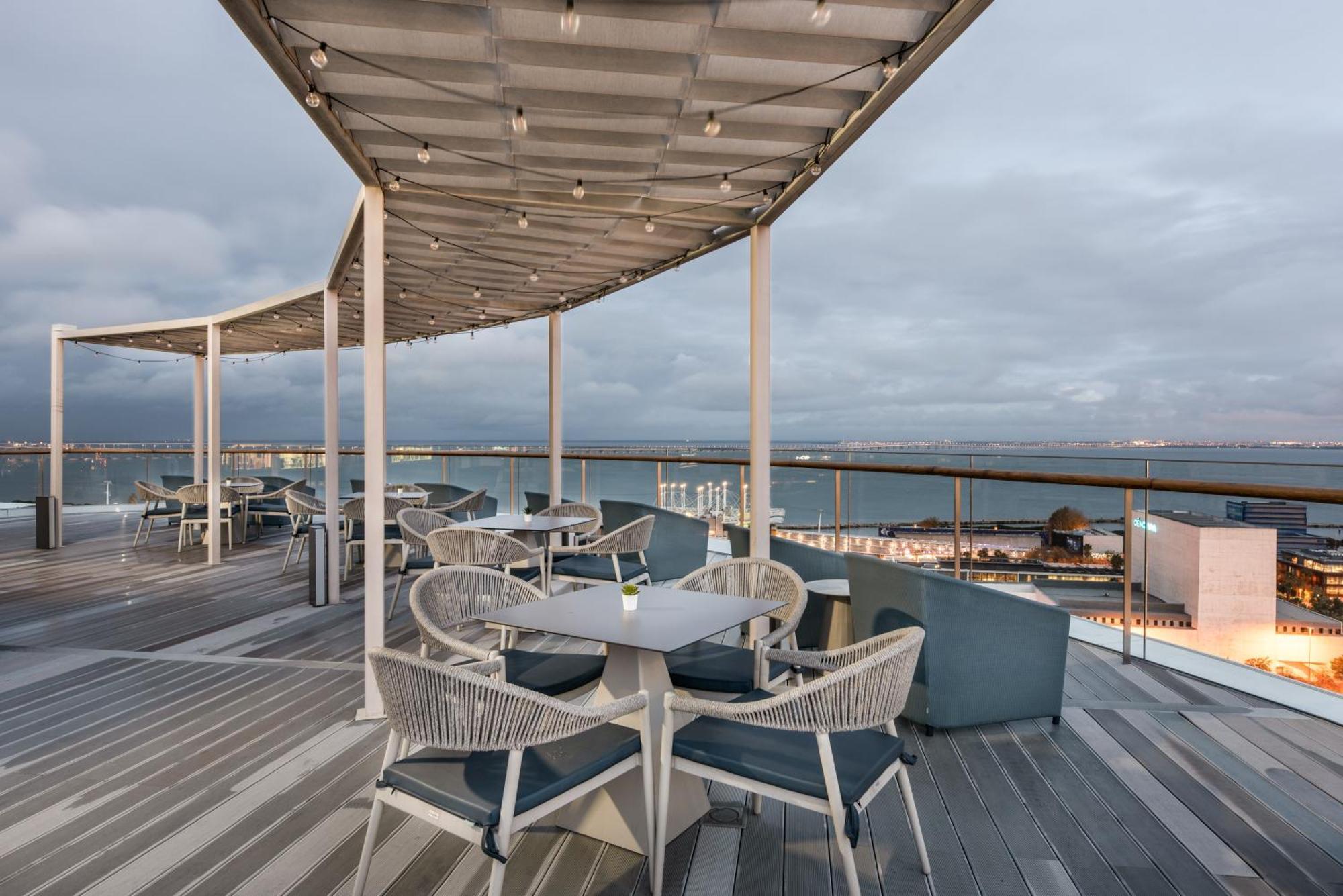 Отель Eurostars Universal Lisboa Экстерьер фото The photo shows a scenic outdoor dining area with modern furniture. There are several round tables and chairs arranged on a wooden deck. Some of the tables have blue cushions, and the area is covered by a sleek, curved canopy that is adorned with str