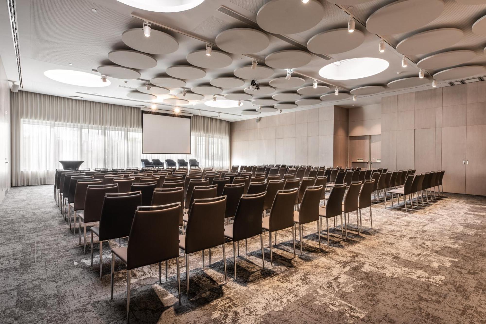 Отель Eurostars Universal Lisboa Экстерьер фото The photo shows a conference or meeting room that is set up for an event. There are rows of brown chairs arranged facing a screen at the front of the room. The space has a modern design, with a decorative ceiling featuring circular elements. Large wi