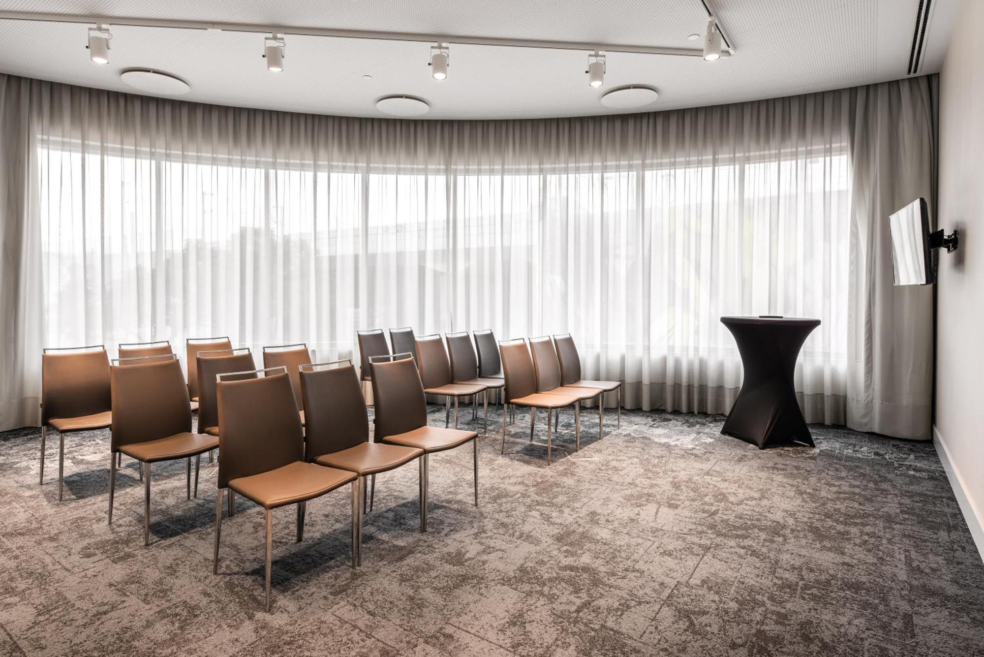 Отель Eurostars Universal Lisboa Экстерьер фото The photo shows a modern meeting room with a curved design. There are several rows of simple, brown chairs arranged in the space, facing a small black podium. The room features large windows covered with sheer curtains, allowing natural light to filt