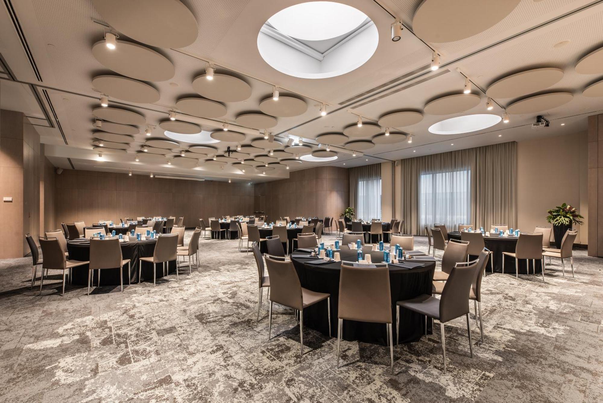 Отель Eurostars Universal Lisboa Экстерьер фото The photo shows a spacious banquet room or conference hall. There are several round tables set up, each covered with black tablecloths and featuring light blue decorations or tableware. The room has a modern design, with a beige and soft gray color p