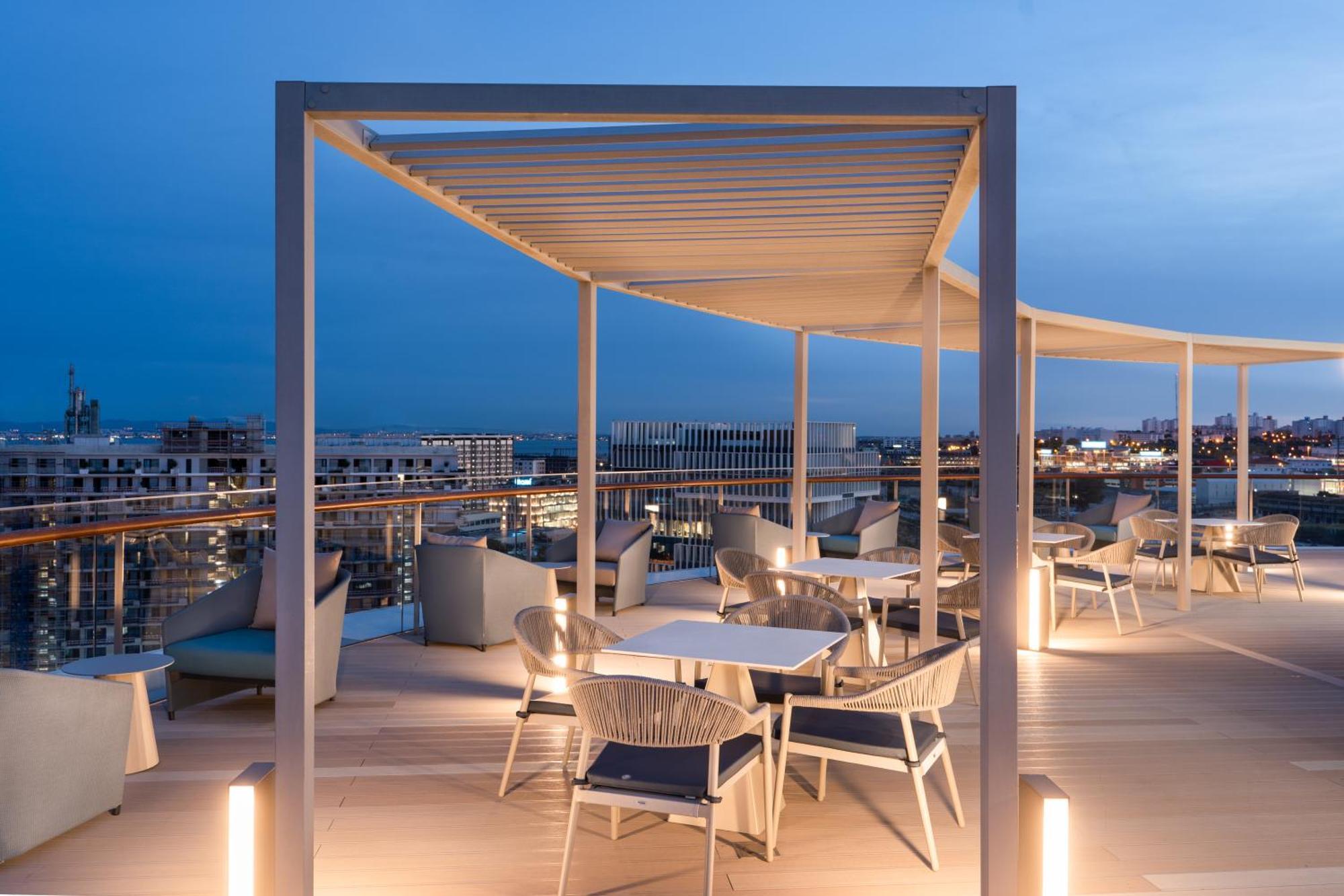 Отель Eurostars Universal Lisboa Экстерьер фото The photo shows a stylish rooftop terrace area, likely part of a restaurant or lounge. It features modern outdoor furniture, including tables and chairs, arranged on a wooden deck. A white pergola provides shade overhead, and there are lights enhanci