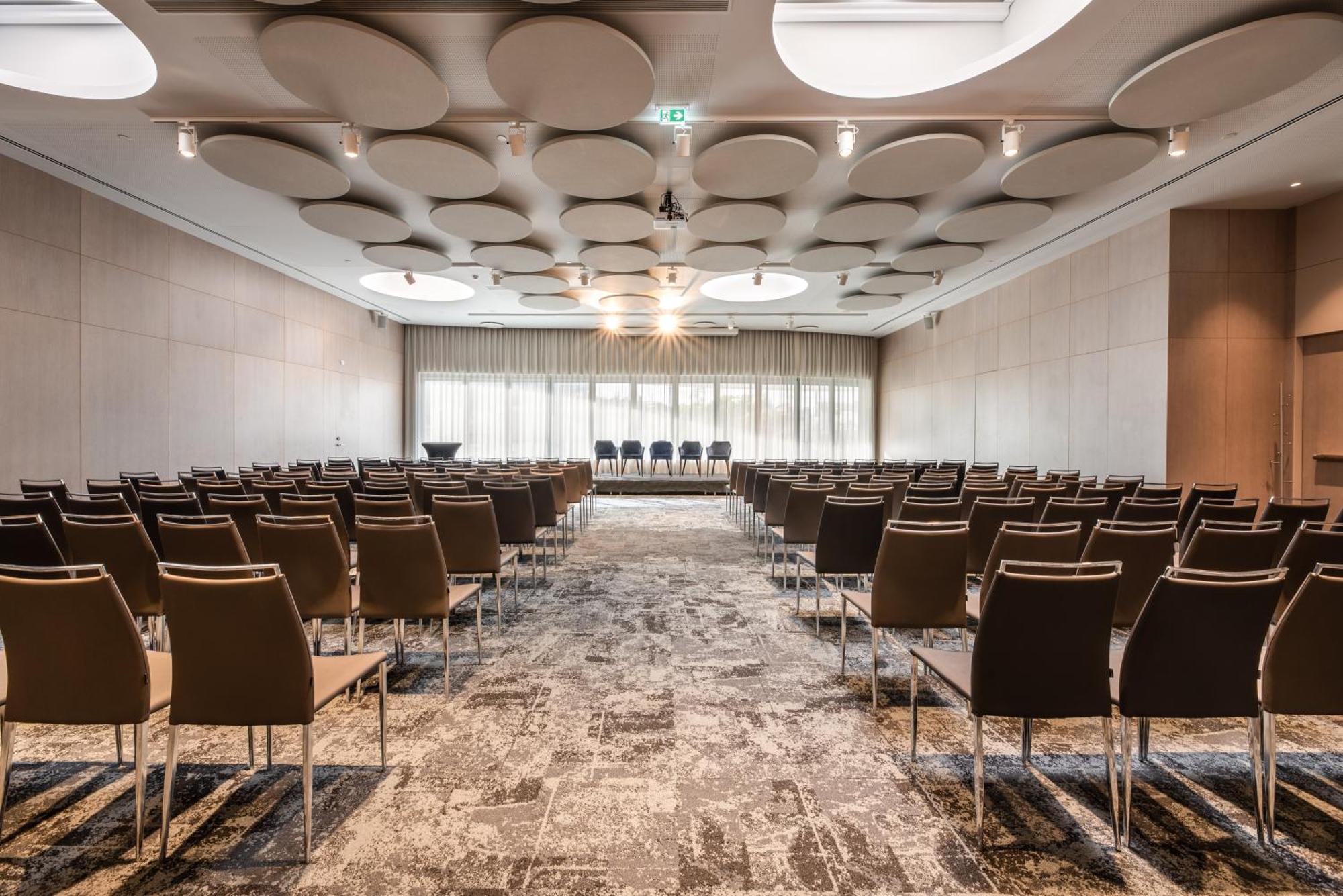 Отель Eurostars Universal Lisboa Экстерьер фото The photo shows a conference or meeting room set up for an event. The room features rows of brown chairs arranged facing a small stage or presentation area at the front. The ceiling has a unique design with circular panels, adding an interesting arch